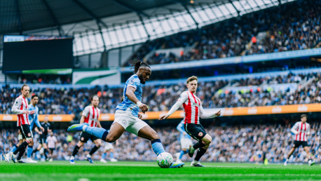 A soccer match with a player in a light blue jersey preparing to kick the ball, surrounded by players in red and white striped jerseys, inside a stadium filled with spectators.
