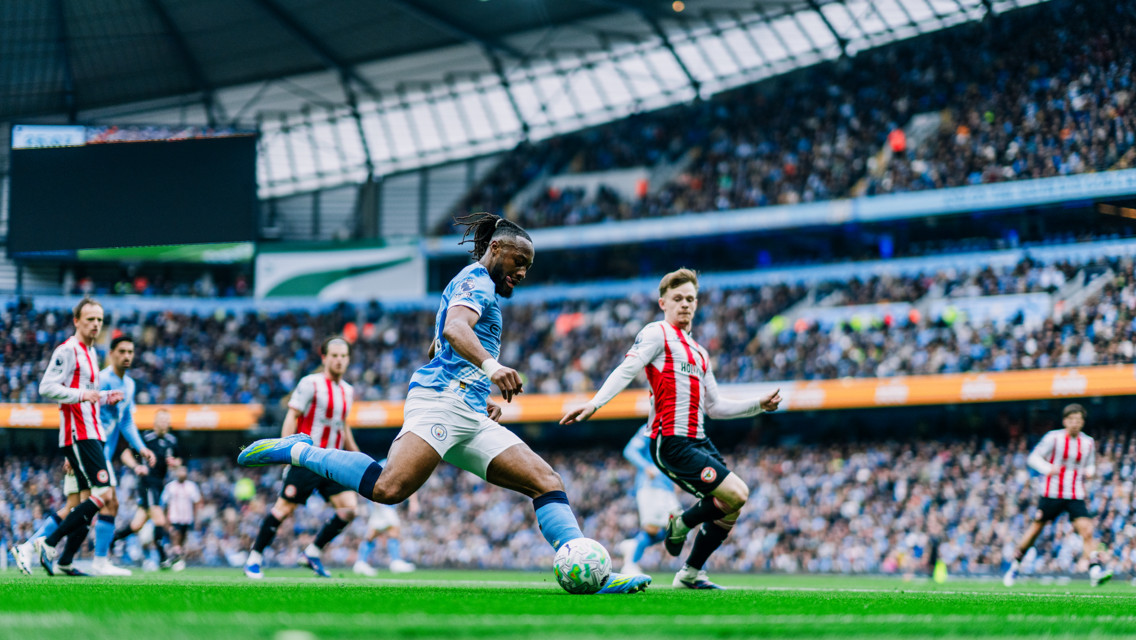 A soccer match with a player in a light blue jersey preparing to kick the ball, surrounded by players in red and white striped jerseys, inside a stadium filled with spectators.