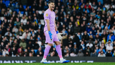 A football player wearing a pink goalkeeper kit and blue gloves is shouting on the field, with a crowd in the background.