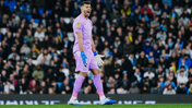 A football player wearing a pink goalkeeper kit and blue gloves is shouting on the field, with a crowd in the background.