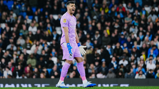 A football player wearing a pink goalkeeper kit and blue gloves is shouting on the field, with a crowd in the background.