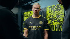 A person wearing a black Manchester City uniform with yellow accents stands in front of posters featuring a bee and text: 'Hive' and 'State of Mind'.