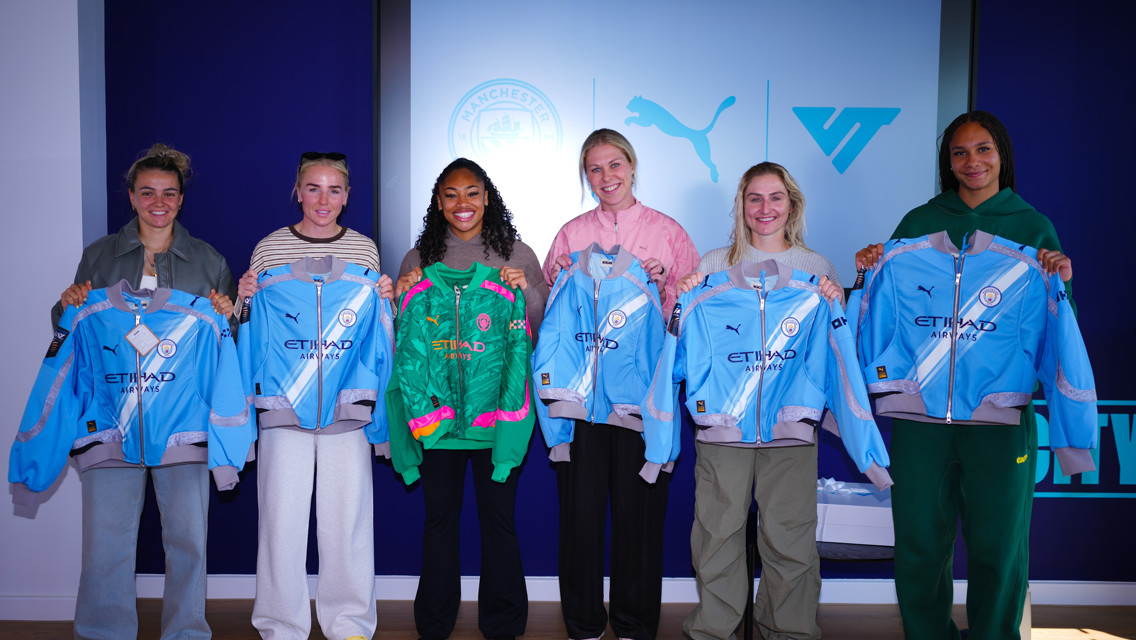 A group of six people holding Manchester City jackets in front of a screen displaying logos of Manchester City and Puma.
