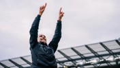 A person in a black jacket raises both arms in celebration at a stadium.
