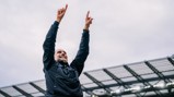 A person in a black jacket raises both arms in celebration at a stadium.