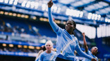 Women soccer players celebrating in light blue jerseys, one with a fist raised, in a stadium.