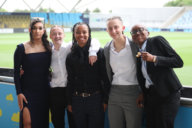 A group of five young individuals dressed formally, posing at an outdoor stadium with blurred faces.