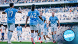 Manchester City players celebrating a goal at Etihad Stadium, wearing light blue kits with crowd in background.