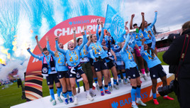 A women's soccer team in blue jerseys celebrating on a podium with fireworks and a Champion banner in the background.