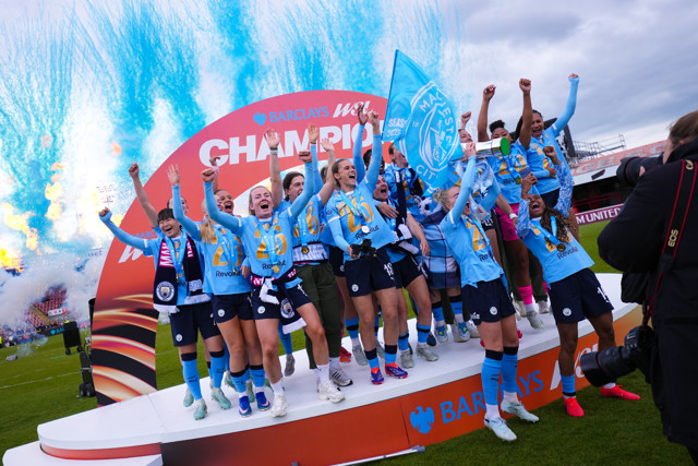 A women's soccer team in blue jerseys celebrating on a podium with fireworks and a Champion banner in the background.