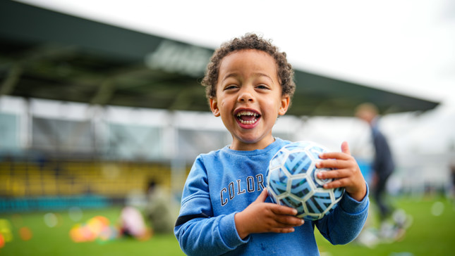 A young child with a blurred face is holding a soccer ball and standing on a soccer field in front of a stadium.