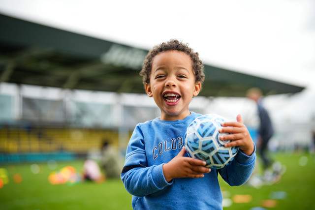 A young child with a blurred face is holding a soccer ball and standing on a soccer field in front of a stadium.