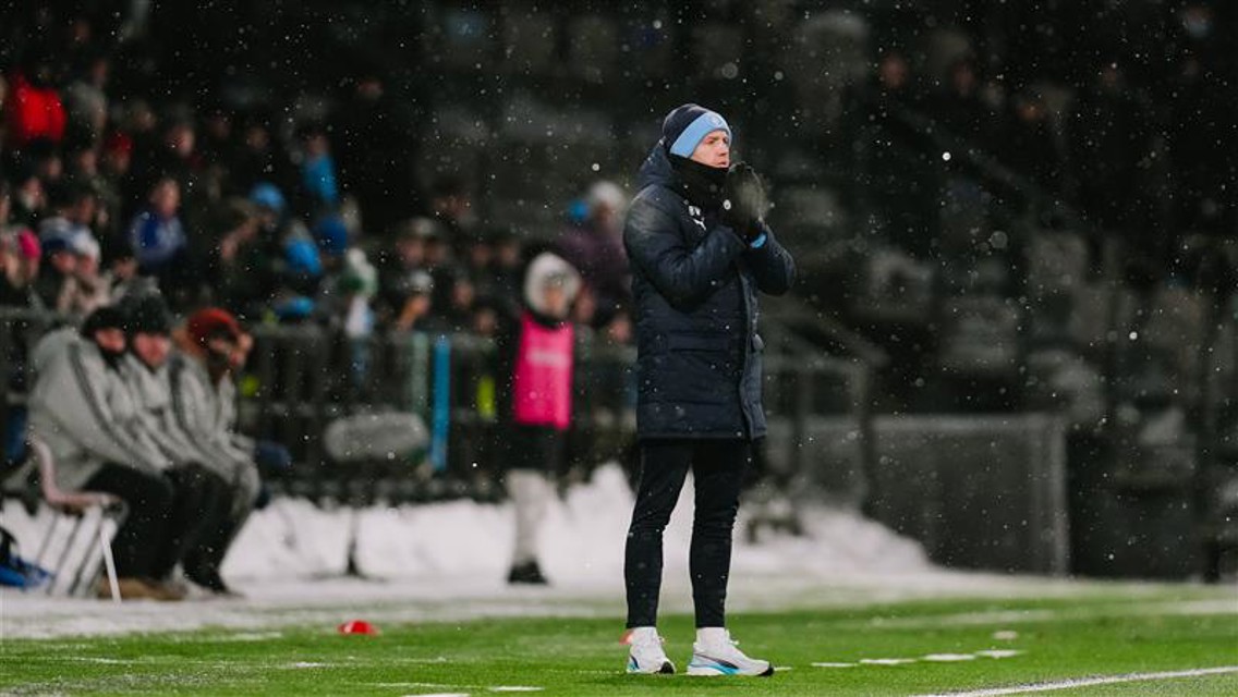 A person in a winter coat stands on the sideline of a soccer field during a snowy match. Snow is visible on the ground and in the air. The stadium is filled with spectators.