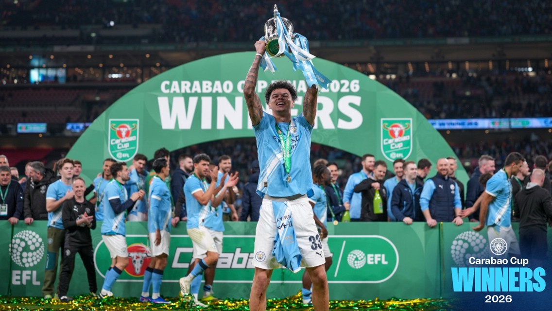 A football player in a light blue kit holds up a trophy surrounded by his team celebrating the Carabao Cup victory in 2026.