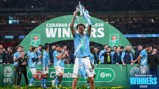 A football player in a light blue kit holds up a trophy surrounded by his team celebrating the Carabao Cup victory in 2026.