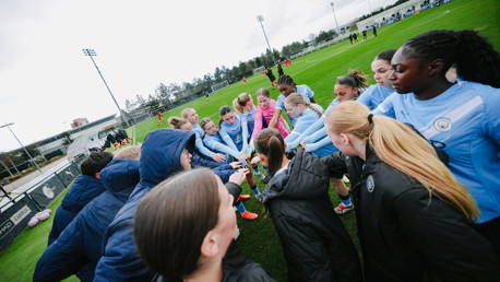A group of female soccer players in light blue jerseys huddle together, putting their hands in the center, on a grassy field with goal posts visible in the background.
