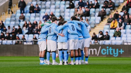 A soccer team wearing light blue jerseys and white shorts huddles together on a field with spectators in the background.