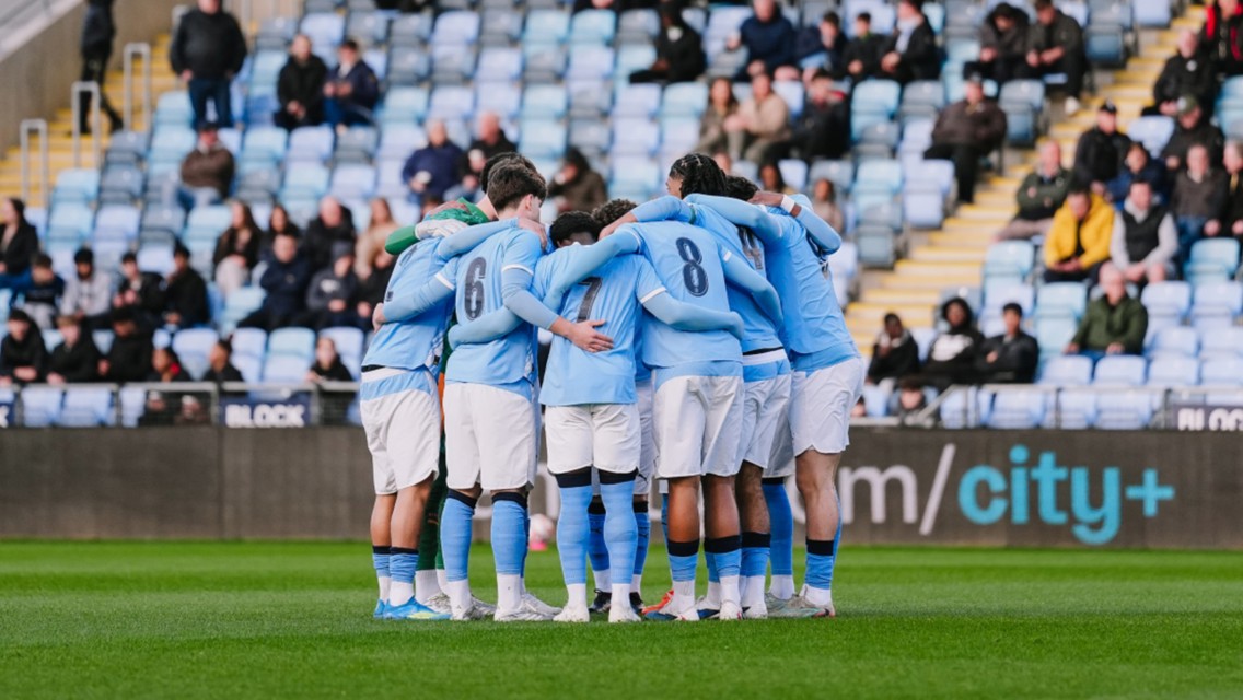 A soccer team wearing light blue jerseys and white shorts huddles together on a field with spectators in the background.