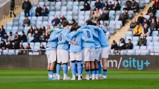 A soccer team wearing light blue jerseys and white shorts huddles together on a field with spectators in the background.