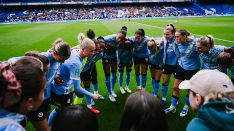 A team huddle of Manchester City women football players in blue jerseys on a soccer field before a match with a stadium crowd in the background.