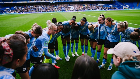 A team huddle of Manchester City women football players in blue jerseys on a soccer field before a match with a stadium crowd in the background.
