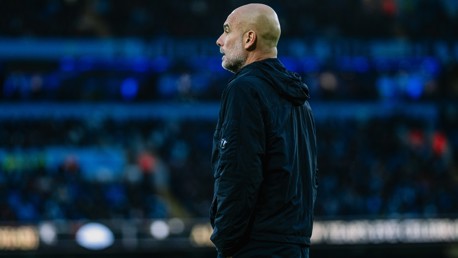 A bald man, likely a coach, wearing a dark jacket observing a football match. The background is a blurred crowded stadium with lights visible.
