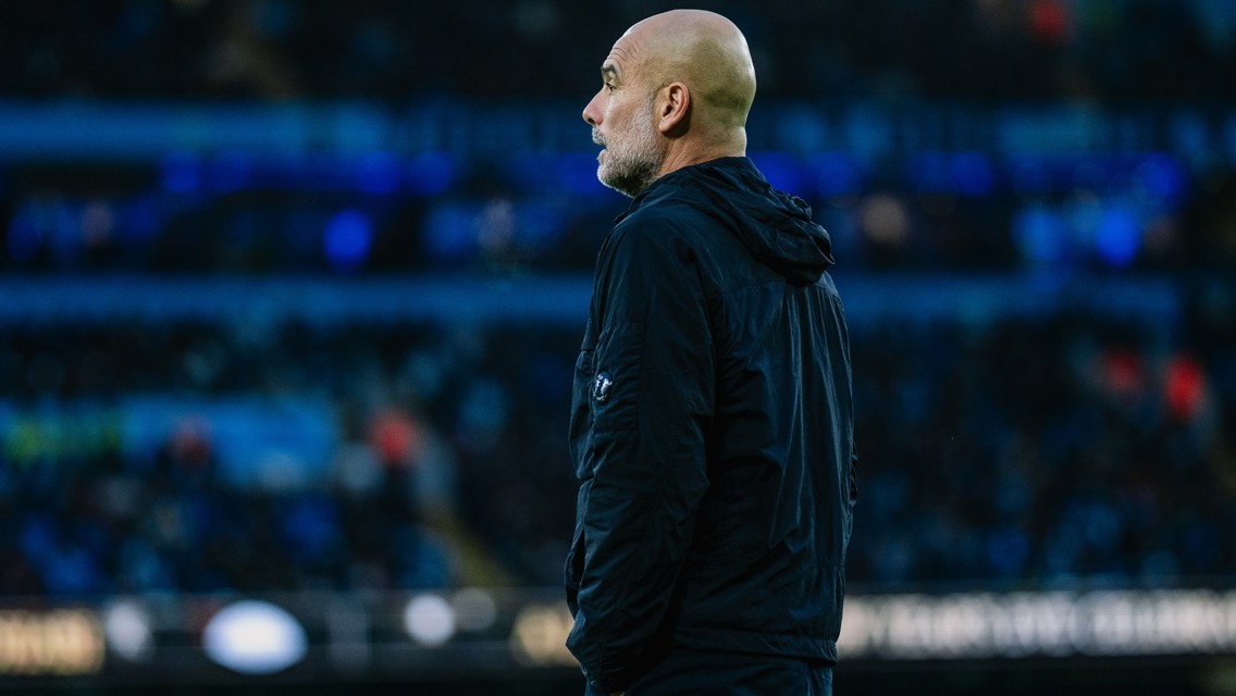A bald man, likely a coach, wearing a dark jacket observing a football match. The background is a blurred crowded stadium with lights visible.