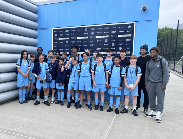 A group of young individuals in Manchester City football attire posing for a team photo outside a building with branding related to Manchester City Football Academy.