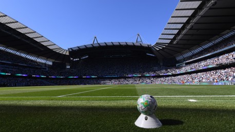 Panoramic view of a soccer stadium with a ball on the field and a large crowd in the stands. The stadium features a distinctive roof structure and clear blue sky.