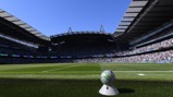 Panoramic view of a soccer stadium with a ball on the field and a large crowd in the stands. The stadium features a distinctive roof structure and clear blue sky.