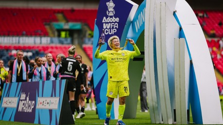 A football player in yellow kit celebrating with a trophy at the Adobe WFA Cup event. Other players are seen wearing medals and scarves.