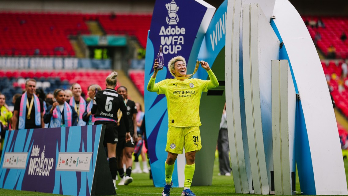 A football player in yellow kit celebrating with a trophy at the Adobe WFA Cup event. Other players are seen wearing medals and scarves.