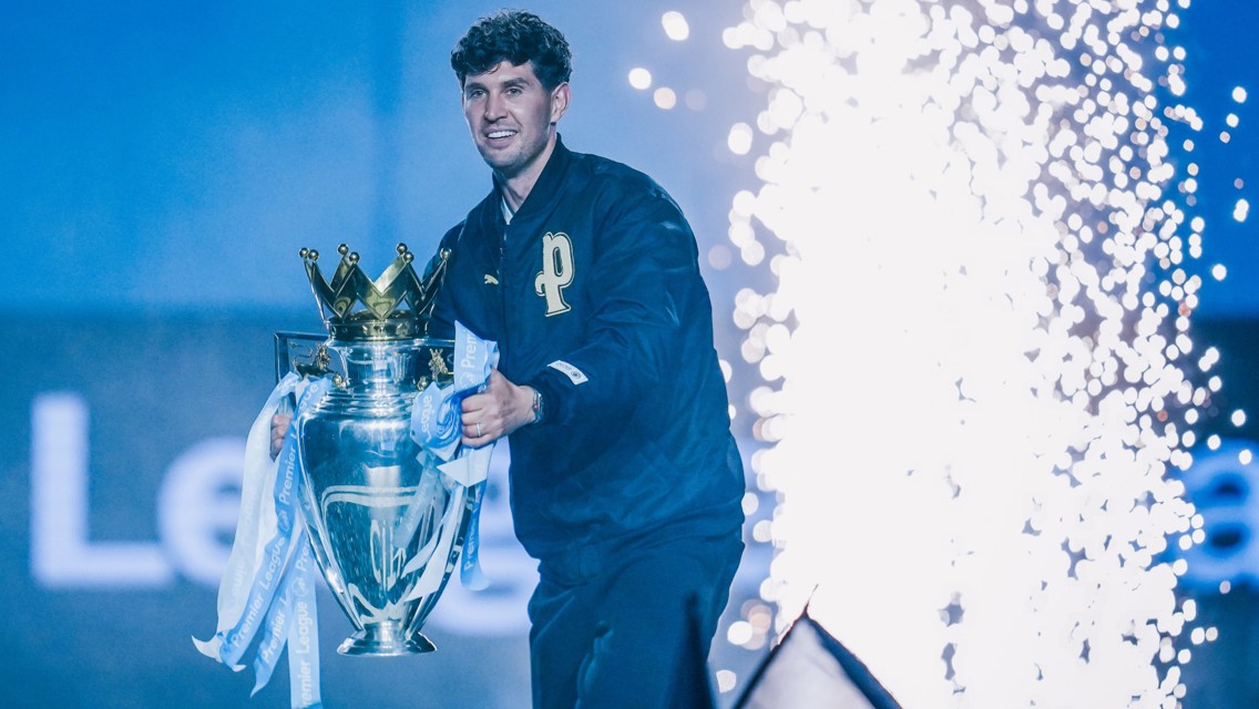 A person holding the Premier League trophy with fireworks in the background, wearing a jacket with a 'P' emblem.