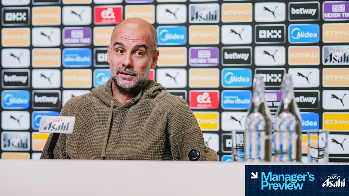 A man in a hoodie sits at a press conference table at Etihad Stadium with Manchester City sponsor logos in the background. Two bottles of water are on the table. The image is part of a manager's preview.