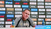 A man at a press conference with an Asahi sign, sitting in front of a backdrop featuring logos like Barclays and Betway. Two Lucozade bottles are on the table labeled 'Manager's Preview.'