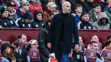 A bald man wearing a dark jacket stands on the sideline during a sports match. He is surrounded by spectators seated in the background.