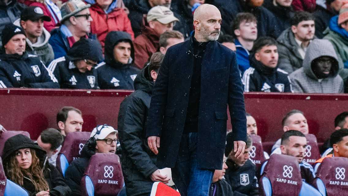 A bald man wearing a dark jacket stands on the sideline during a sports match. He is surrounded by spectators seated in the background.