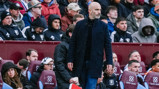 A bald man wearing a dark jacket stands on the sideline during a sports match. He is surrounded by spectators seated in the background.