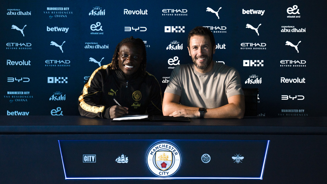 Two individuals at a table sign a document with Manchester City branding in the background.