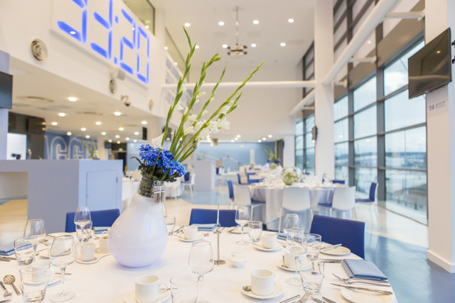 An elegant event venue with round tables set for a formal occasion. The room features large windows, white tablecloths, blue chairs, white and blue floral centerpieces, and a modern decor. A large digital clock is mounted on the wall.
