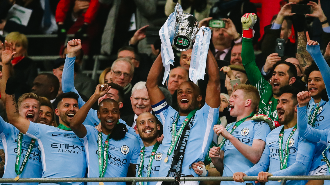 Football players in light blue kits lifting a trophy with confetti in the air.
