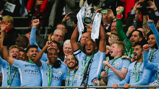 Football players in light blue kits lifting a trophy with confetti in the air.