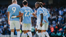 Four Manchester City players celebrating on the pitch wearing light blue jerseys with names and numbers on their backs. The crowd is visible in the background.