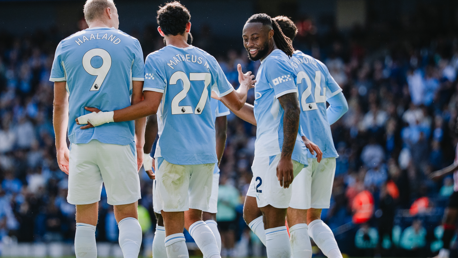 Four Manchester City players celebrating on the pitch wearing light blue jerseys with names and numbers on their backs. The crowd is visible in the background.