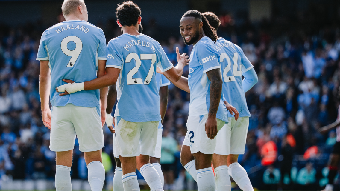 Four Manchester City players celebrating on the pitch wearing light blue jerseys with names and numbers on their backs. The crowd is visible in the background.