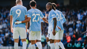Four Manchester City players celebrating on the pitch wearing light blue jerseys with names and numbers on their backs. The crowd is visible in the background.