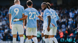 Four Manchester City players celebrating on the pitch wearing light blue jerseys with names and numbers on their backs. The crowd is visible in the background.