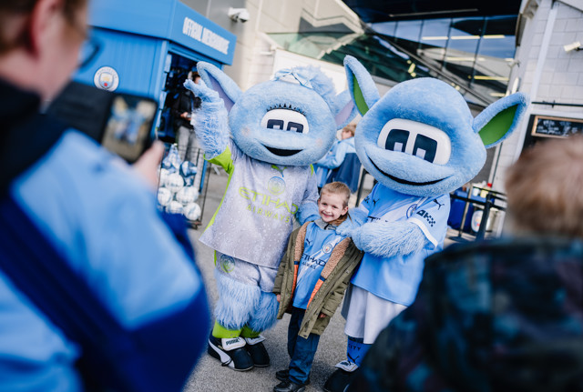 A child poses with two Manchester City mascots, Moonbeam and Moonchester, in front of the Manchester City official merchandise store.
