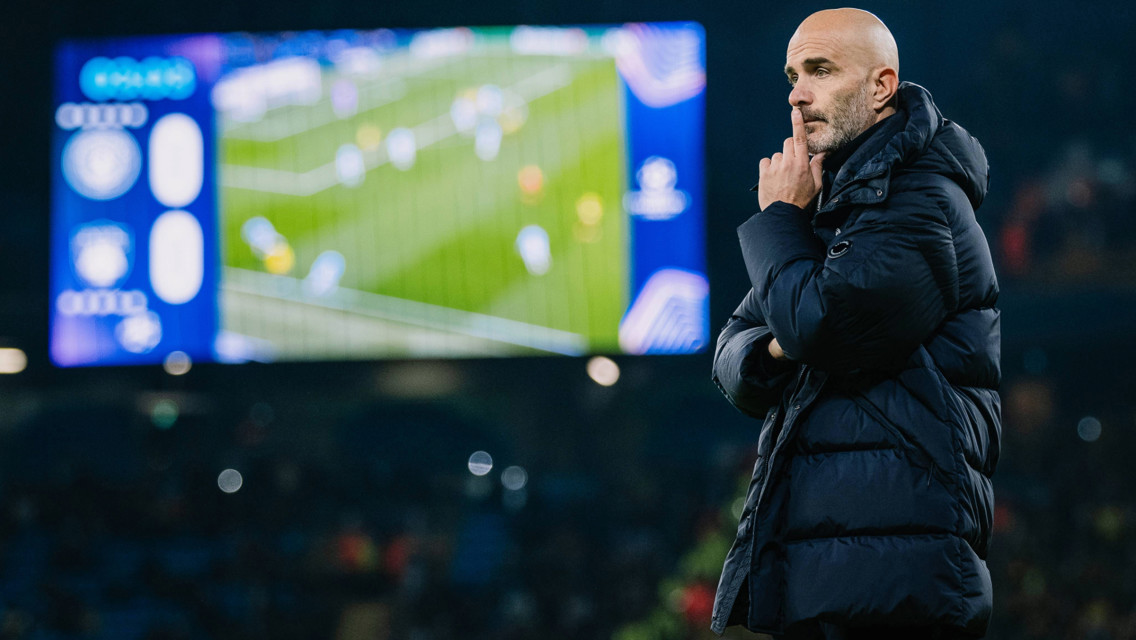 A soccer coach wearing a black jacket stands on the sideline, holding his chin and watching a match. A blurred screen in the background shows the game with scoreboards.
