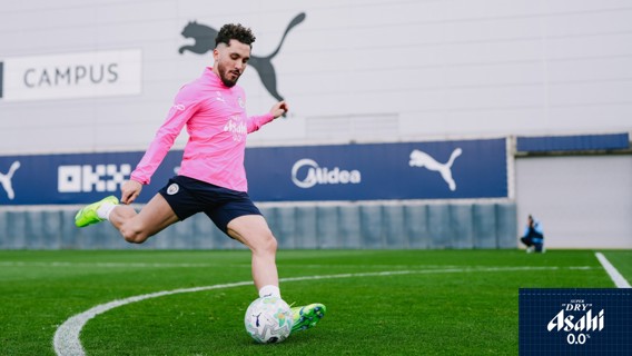 A football player in a pink training kit kicking a ball on a green pitch with Puma branding in the background.